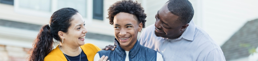 A foster care involved family smiles in front of their home. A foster mom and foster dad have their hands rested reassuringly on their teen foster son's shoulder as they smile together.
